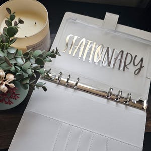 May include: A white binder with the word "STATIONARY" in silver lettering. The binder has a clear pocket and silver rings. A lit candle and a small plant are in the background.