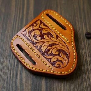 Op de afbeelding: Een handgemaakte, bruin lederen holster met een bloemmotief. De holster heeft een lichtbruine rand met gele stiksels en twee sleuven. Een koffieboon ligt op het houten oppervlak.
