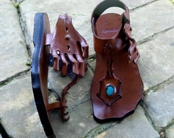 May include: Handmade brown leather sandals with unique hand-shaped accents. The sandals feature a turquoise stone embellishment and a black sole. The design includes intricate cutouts and a strap for the ankle.