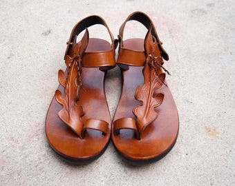 May include: A pair of brown leather sandals with a leaf design on the front. The sandals have a toe loop and ankle strap. The sandals are on a light gray surface.