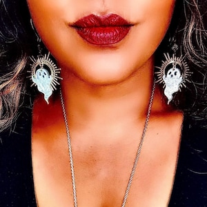 May include: A pair of silver earrings with a ghost design and a sunburst halo. The earrings are hanging from the ears of a person with dark hair and red lipstick.