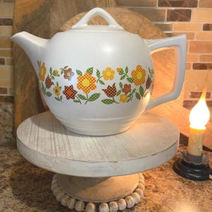 May include: White ceramic teapot with a floral pattern in orange, yellow, and brown. The teapot has a handle and a spout, and sits on a white wooden stand. The stand is decorated with a beaded trim.