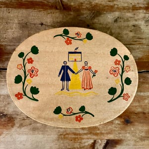 May include: Oval wooden serving tray with a painted folk art design. The design features a couple holding hands in front of a building, surrounded by floral vines. The tray is light brown with red, yellow, green, and blue accents.
