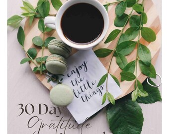 Può includere: Un diario di gratitudine di 30 giorni con il testo "enjoy the little things" su un biglietto bianco con macarons verdi e una tazza di caffè su un tagliere di legno con foglie verdi.