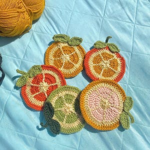 May include: Five crocheted orange slice coasters with green leaves. The coasters are made with a variety of colors, including orange, yellow, green, and pink.