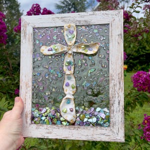 May include: A framed, handcrafted cross made with oyster shells, iridescent glass, and colorful gems. The cross is set against a shimmering background and framed in a distressed white wooden frame. The artwork is displayed outdoors, surrounded by greenery and flowers.
