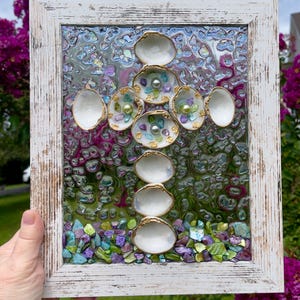 May include: A framed cross-shaped artwork featuring white seashells with gold trim, arranged on a textured glass background. The glass is filled with colorful crushed stones and small pearls. The frame is white with a distressed finish.