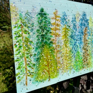 K&ouml;nnte beinhalten: Ein Gem&auml;lde, das eine Waldszene mit B&auml;umen aus gr&uuml;nen, blauen und goldenen Glasst&uuml;cken zeigt. Der Hintergrund ist hellblau mit einer schimmernden Textur.