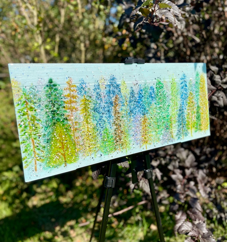 K&ouml;nnte beinhalten: Ein rechteckiges Gem&auml;lde auf einem Holzstaffelei zeigt eine Waldszene in einem blauen, gr&uuml;nen und goldenen Farbschema. Die B&auml;ume bestehen aus kleinen, schillernden Glasst&uuml;cken, die einen funkelnden Effekt erzeugen.