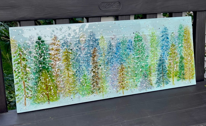 K&ouml;nnte beinhalten: Ein Glaskunstwerk, das eine Waldszene mit einer Vielzahl von blauen, gr&uuml;nen und goldenen B&auml;umen darstellt. Der Hintergrund ist hellblau mit einem subtilen Glitzereffekt.