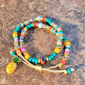 May include: A colourful beaded bracelet with a pineapple charm. The bracelet is made of turquoise, yellow, pink, orange, and white beads. The pineapple charm is gold and has a green top.