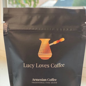 May include: A black resealable bag of Armenian coffee with a gold illustration of a traditional coffee pot. The bag reads "Lucy Loves Coffee" and "Armenian Coffee Traditional Fine Grind" with a net weight of 1 ounce (28 grams).