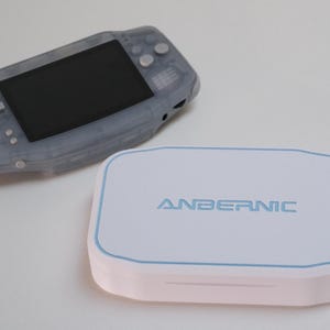May include: A handheld gaming console with a translucent grey plastic shell and a black screen, positioned next to a white rectangular storage case featuring the blue text ANBERNIC and a thin blue border outline on the top surface.