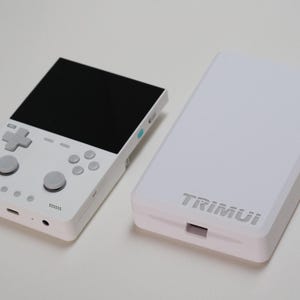 May include: A white handheld gaming console with a black screen, grey directional pad, two analogue sticks, and four action buttons, positioned next to a matching white rectangular protective case with the word TRIMUI embossed on the front surface.