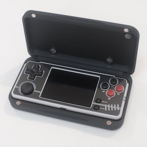 May include: A black handheld games console with a silver and black faceplate. The console features a directional pad, a joystick, and red and black buttons. It is housed in a black case with a magnetic closure.