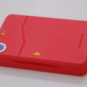 May include: A rectangular red plastic case with a textured surface, featuring a circular blue button with a white border, three small circular indicator lights in green, yellow, and red, and a small yellow triangle icon printed on the front.