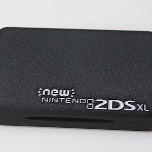 Op de afbeelding: Een zwarte draagbare spelcomputer met een getextureerde matte afwerking. De bovenkant is voorzien van de witte reliëftekst "new NINTENDO 2DS XL" in het midden onderaan. Het apparaat heeft een rechthoekige vorm met afgeronde hoeken en een slank profiel.