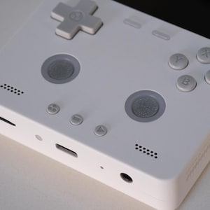 May include: A white handheld video game console featuring a directional pad, two circular analog sticks, and four action buttons labeled X, Y, A, and B. The device includes a speaker grille, a headphone jack, and a USB-C port on the bottom edge.