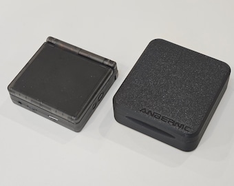 May include: A black handheld gaming console with a flip-up screen is next to a black textured case with the word "ANBERNIC" embossed on it. The console is open, revealing the screen and buttons.