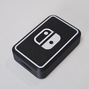 May include: Black rectangular case with a white outline and a white graphic of a game controller. The case has a textured surface and a small opening on one side. The case is likely for a gaming console.