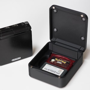 May include: Black Nintendo Game Boy Advance SP console next to a black case holding two game cartridges. One cartridge is red with "Pokemon Ruby Version" text. The other cartridge is white with "Final Fantasy Tactics" text.