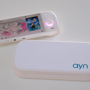 Könnte beinhalten: Eine weiße tragbare Spielkonsole mit einem Bildschirm, der ein Spielmenü anzeigt, und leuchtenden blauen und rosa Joysticks. Daneben befindet sich eine passende weiße rechteckige Schutzhülle mit dem Wort ayn in blaugrüner Schrift.