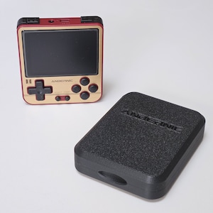 May include: A handheld retro gaming console with a gold and burgundy color scheme, featuring a screen, directional pad, and buttons. A black textured case with the brand name "ANBERNIC" is also present.