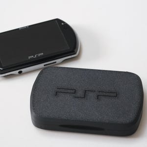 Może przedstawiać: Czarna i srebrna konsola do gier przenośnych Sony PSP z czarnym ekranem, obok czarnej obudowy z logo PSP. Konsola znajduje się na białej powierzchni.