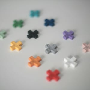 Può includere: Un assortimento di blocchi di costruzione a forma di croce colorati, tra cui giallo, arancione, rosso, rosa, bianco, grigio, nero, verde, verde acqua e viola. I blocchi sono sparsi su una superficie bianca.