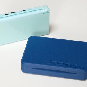 May include: Two Nintendo DS Lite consoles. One is a light blue color, closed, and the other is a dark blue color with the text "NINTENDO DS lite" on the front. The consoles are on a white surface.