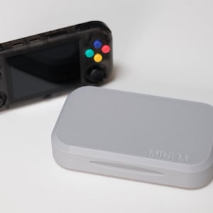 Puede incluir: Una consola de juegos portátil con una carcasa negra transparente, con una pantalla negra, un mando en cruz y botones de colores. Al lado, una funda rectangular gris con el texto "MMMM".