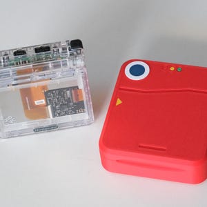 May include: A clear, transparent plastic handheld gaming console shell with visible internal circuitry and a Nintendo logo, positioned next to a solid red, square-shaped electronic device featuring a blue circular button and three small indicator lights.