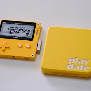 May include: A bright yellow handheld gaming console with a square screen displaying the text Whitewater Wipeout and a matching yellow protective cover featuring the embossed word playdate. The device includes a directional pad and two buttons.