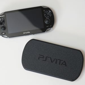 以下が含まれることがあります： 黒いPS Vita携帯ゲーム機と、それに合う黒いケース。コンソールは光沢のある画面、コントロールボタン、Sonyのロゴを備えています。ケースにはPS Vitaのロゴがエンボス加工されています。白い背景に配置されています。