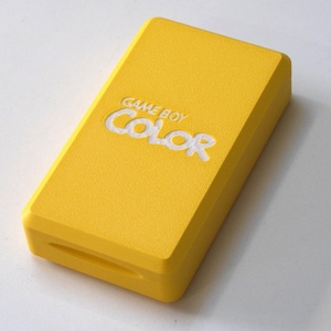 Könnte beinhalten: Eine leuchtend gelbe Game Boy Color-Hülle mit den Worten "GAME BOY COLOR" in weißer Schrift. Die Hülle hat eine strukturierte Oberfläche und eine rechteckige Form mit abgerundeten Kanten. Ein Schlitz ist unten sichtbar.