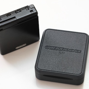 Könnte beinhalten: Schwarze Game Boy Advance SP Konsole und passende Hülle. Die Konsole ist rechteckig mit einem Klappdesign und einem Bildschirm. Die Hülle ist quadratisch mit den Worten "GAME BOY ADVANCE SP" auf der Oberseite geprägt. Das Nintendo-Logo ist auf der Konsole sichtbar.