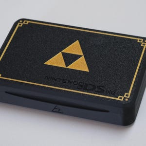 Puede incluir: Una consola de juegos portátil Nintendo DS Lite negra con un emblema Triforce dorado en la cubierta superior. El dispositivo incluye un diseño de borde rectangular dorado y el texto en relieve NINTENDO DS LITE debajo del símbolo triangular central.