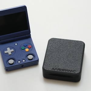 May include: A navy blue handheld gaming console with a black screen, open, and a gray directional pad and colorful buttons. Next to it is a black, textured case with the word "ANBERNIC" printed on it.