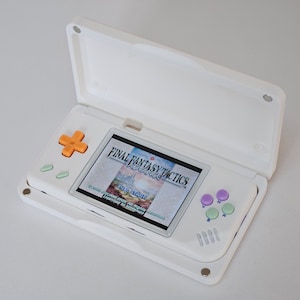 May include: White handheld gaming console with an open lid, displaying a game screen. The console features orange, green, and purple buttons. The game on the screen is "FINAL FANTASY TACTICS ADVANCE".