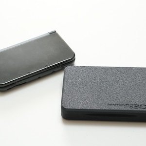 Könnte beinhalten: Zwei schwarze Nintendo 3DS-Konsolen werden auf einer weißen Oberfläche gezeigt. Eine ist geöffnet und zeigt die Bildschirme, während die andere ein strukturiertes Gehäuse mit dem Text "NINTENDO 3DS" ist. Diese Konsolen sind für Handheld-Gaming konzipiert.