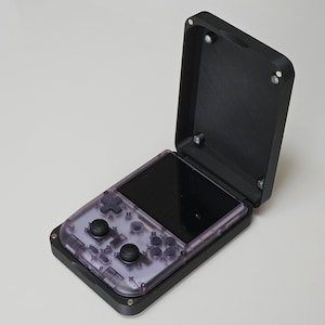 Op de afbeelding: Een handheld gameconsole met een transparante paarse behuizing en zwarte bedieningselementen, gehuisvest in een zwarte beschermhoes. De hoes is open en onthult het scherm en de knoppen. Het apparaat is ontworpen voor draagbaar gamen.