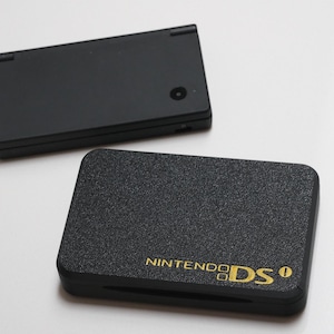 May include: A black handheld gaming console with a smooth finish and a camera lens on the top cover, positioned above a black textured protective case featuring the gold embossed text NINTENDO DSi on the lower right corner of the front surface.