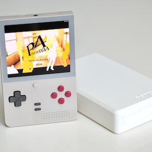 Könnte beinhalten: Ein weißes Handheld-Spielgerät mit einem Bildschirm, auf dem ein Videospiel angezeigt wird. Das Gerät hat ein Steuerkreuz, rosa Aktionstasten und einen Lautsprecher. Daneben befindet sich ein weißes rechteckiges Gehäuse mit dem Wort "RETROID".