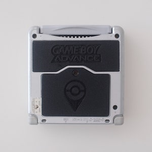 May include: A silver Game Boy Advance handheld game console. The back features a black panel with the words "GAME BOY ADVANCE" embossed. Below the text is a black graphic of a location pin with a Poké Ball symbol.