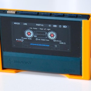 May include: A teal and orange digital music player with a screen showing a cassette tape interface. The player has the text "SNOWSKY" and "DCRO" printed on it. The screen displays the song "City Ruins - Rays of Light".