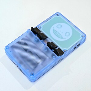 May include: A light blue handheld gaming device with a transparent case. The screen displays a white smiling face graphic. The device has black buttons and a ribbed design on the bottom half.