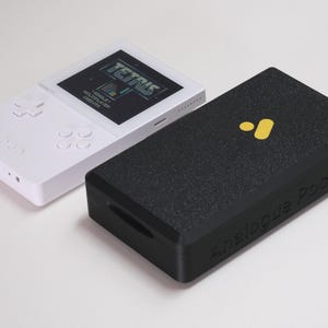 May include: A white handheld gaming console displaying the Tetris game title on its screen, positioned next to a black rectangular protective case with a yellow logo and the embossed text Analogue Pocket on the side of the device.