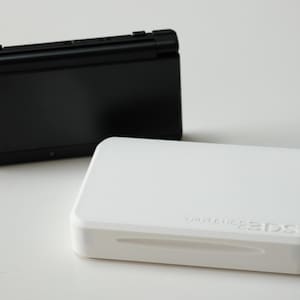 May include: Two Nintendo 3DS consoles, one black and one white, are displayed. The white console has the words "Nintendo 3DS" embossed on its surface. The black console is behind the white one. Both are closed and on a white surface.