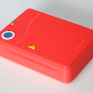 Könnte beinhalten: Ein rotes rechteckiges Gehäuse mit einem blau-weißen Kreis, einem gelben Dreieck und drei kleinen farbigen Kreisen. Das Gehäuse hat eine strukturierte Oberfläche und eine rechteckige Form. Das Gehäuse ist wahrscheinlich ein Aufbewahrungsbehälter oder ein dekorativer Gegenstand.