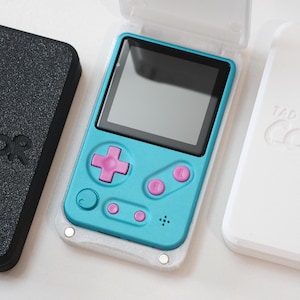 May include: A handheld gaming device with a teal body, pink buttons, and a black screen. The device is in a clear case, with a black case and a white case nearby. The cases and the device all have the text "Tad Boy Color".